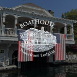 A white waterfront lodge with American flags hanging from its balcony reads “Boathouse Waterfront Lodging.”